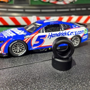 May include: A blue, white, and red race car with the number 5 and the text "HendrickCars.com" is on a track. Two black rubber tires are in the foreground. The car has a detailed design and is positioned on a track with red and white barriers.