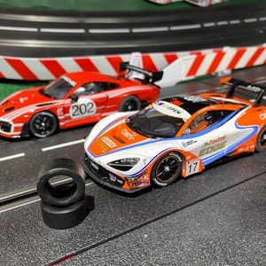 May include: Two slot car race cars on a track. The car in the lead is orange, white, and blue with the number 17 on the side. The car in the back is red with the number 202 on the side. There are two black tires on the track.