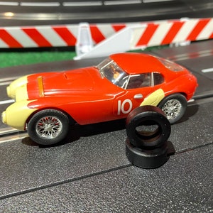 May include: A red and cream-colored toy race car with the number 10 on the side, sitting on a black track. Two black tires are in front of the car. The car has silver wheels and a clear windshield.