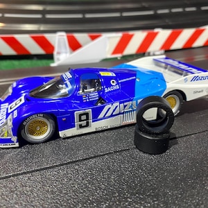 Puede incluir: Un coche de carreras de juguete azul y blanco con el número 9 en el lateral, ruedas doradas y un juego de neumáticos negros. El coche tiene logotipos de patrocinadores y está en una pista negra.