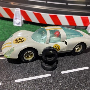 May include: A white toy race car with a gold number 33 on the side. The car is on a black race track with white lines. There are four black tires stacked next to the car.