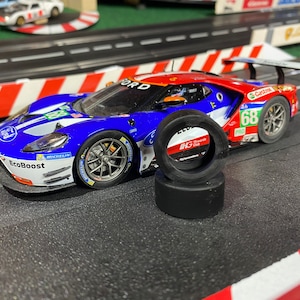 May include: A detailed image of a Ford GT race car model in blue, white, and red, with the number 68. Two black tires are in front of the car. The car is on a black track with red and white barriers.