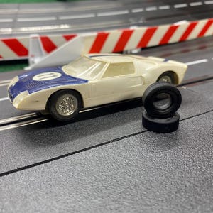May include: A vintage toy race car in cream and blue, with a racing stripe and the number 10 on the hood. Two black tires are stacked next to the car on a gray track. The background includes a white and red barrier.