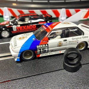 Puede incluir: Un coche de carreras blanco y azul con el número 15 y las palabras "DEKRA" y "WARSTEINER" en el lateral. El coche está en una pista negra con una barrera roja y blanca en el fondo.