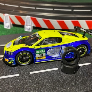May include: A yellow and blue toy race car with the number 44 and the words "DEKRA ADAC" on the side. The car is on a black track with white lines. There are two black tires next to the car.
