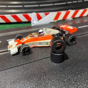 May include: An orange and white race car with black tires. The car is on a black track with a red and white barrier in the background. There are three spare tires stacked on the track.