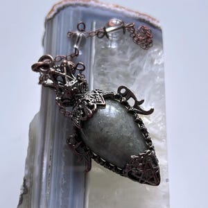 May include: A detailed pendant with a grey, teardrop-shaped gemstone set in dark copper-toned metal. The pendant is suspended from a chain with clear crystal beads. The design has a vintage, gothic aesthetic, with intricate metal detailing.