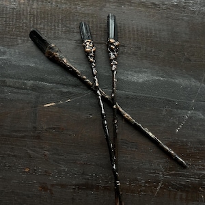 Può includere: Tre bacchette nere e bronzate con punte di cristallo sono esposte su una superficie di legno scuro. Le bacchette presentano intricati dettagli in bronzo e una finitura lucida, suggerendo un tema fantasy o magico.