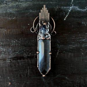 Puede incluir: Colgante artesanal con un cristal azul oscuro engastado en metal color cobre. El colgante tiene un diseño geométrico en la parte superior y está suspendido de una fianza circular. El cristal está facetado y tiene un acabado pulido.