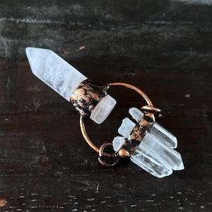 Puede incluir: Un colgante de cristal de cuarzo transparente con detalles metálicos color cobre. El colgante presenta un cristal grande y puntiagudo y grupos de cristales más pequeños, todo ello sujeto por un marco metálico circular. El metal tiene un aspecto texturizado y envejecido.
