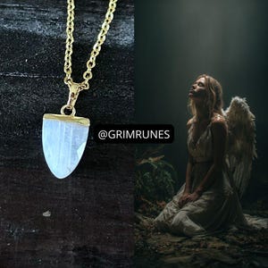 May include: A gold-colored necklace with a pendant featuring a light blue, shield-shaped stone. The necklace is displayed on a dark surface. The image also shows a person with angel wings, wearing a white dress, and looking upwards.