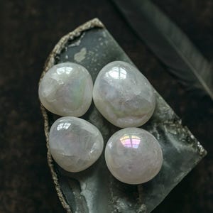 Peut inclure: Quatre pierres polies et iridescentes reposent sur une dalle de pierre grise foncée aux bords rugueux. Les pierres ont un éclat nacré et opalescent, reflétant la lumière avec des reflets arc-en-ciel. Une plume sombre est en arrière-plan.