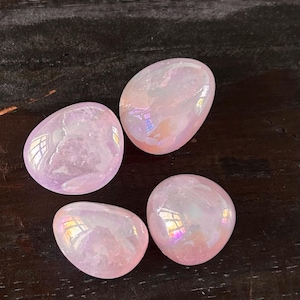 May include: Four polished, oval-shaped stones with an iridescent sheen. The stones are a mix of pale pink and lavender, with flashes of rainbow colours. The stones are smooth and reflective, set against a dark wood surface.