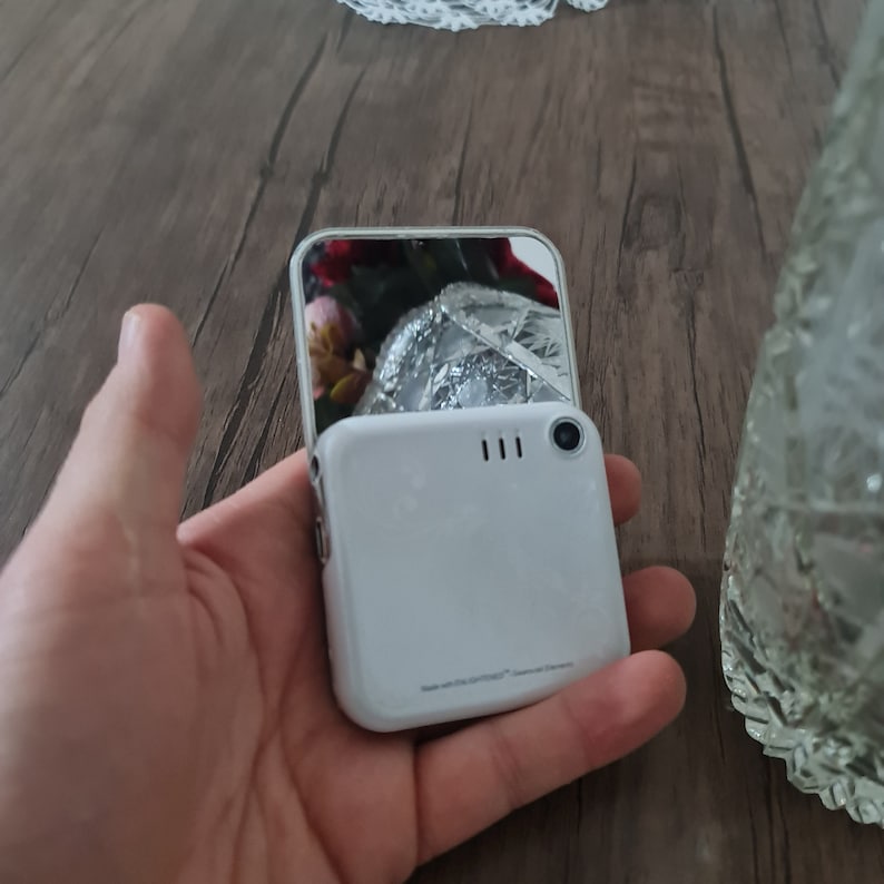 Op de afbeelding: Een witte flip telefoon met een klein scherm aan de voorkant en een groter scherm aan de achterkant. De telefoon is dicht en het achterste scherm toont een reflectie van een bloem.