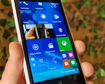 May include: A white Nokia Lumia smartphone with a black screen displaying the Windows Phone operating system. The screen shows various app icons, including "Cumhuriyet", "Mesajlaşma", "Kişiler", "Hava Durumu", "Outlook Posta", "Fotoğraflar", "Groove Müzik", "Filmler ve TV", "Xbox", "Microsoft Edge", "Haritalar", and "Tüm uygulamalar →".