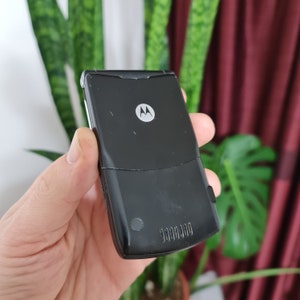 May include: A black Motorola flip phone with a silver Motorola logo on the front. The phone has a small, round button on the bottom.