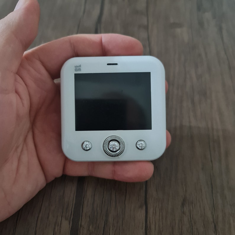 Op de afbeelding: Een witte mobiele telefoon met een zwart scherm en drie zilveren knoppen. De telefoon heeft de tekst "GM" in de linkerbovenhoek.