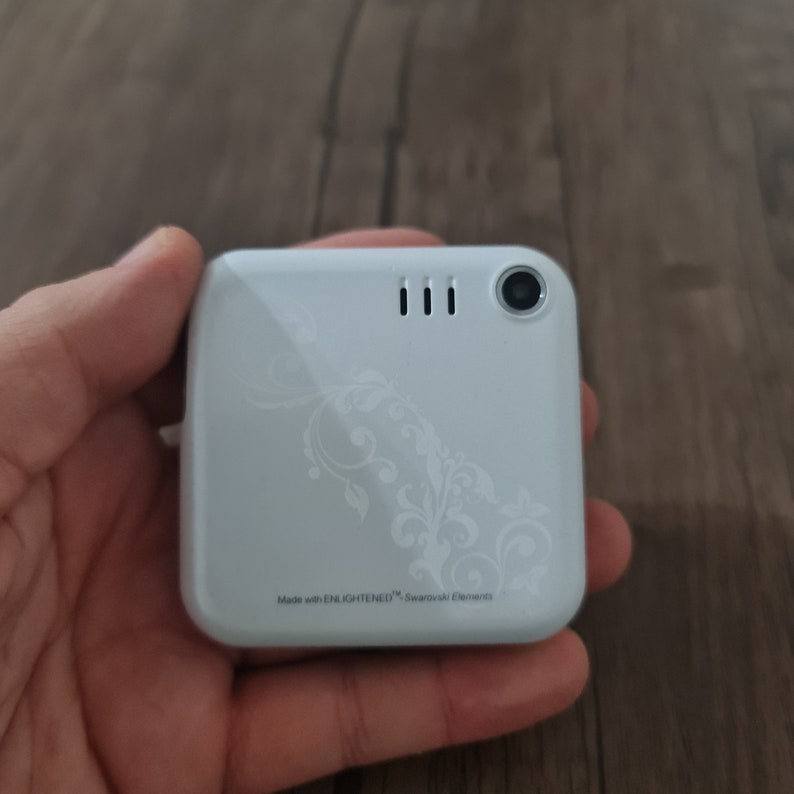Op de afbeelding: Witte mobiele telefoon met een decoratief bloemmotief en de tekst "Made with ENLIGHTENED&trade; Swarovski Elements".