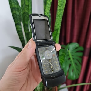 May include: A black Motorola flip phone with a silver trim and a keypad with yellow numbers. The phone is open and the screen is black.