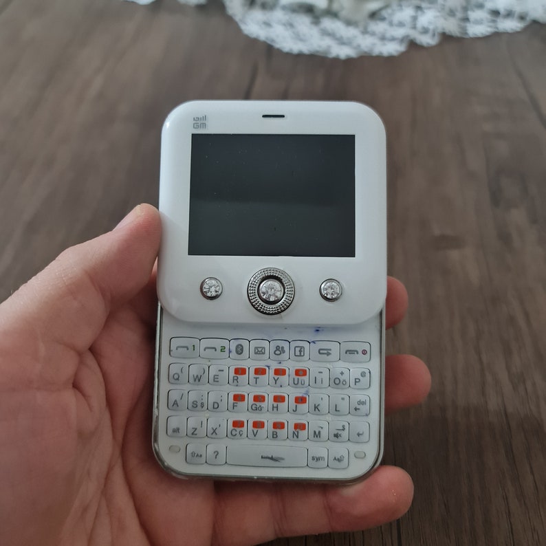 Op de afbeelding: Een witte mobiele telefoon met een klein scherm en een qwerty-toetsenbord. De telefoon heeft een zilveren rand en een paar strasssteentjes aan de voorkant.