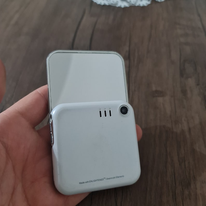 Op de afbeelding: Witte flip telefoon met een camera lens en de tekst "Made with ENLIGHTENED&trade;  Survival Elements" op de achterkant.