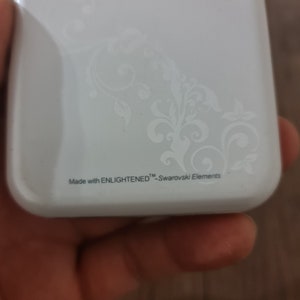 Op de afbeelding: Witte telefoonhoes met een bloemenpatroon en de tekst "Made with ENLIGHTENED&trade; - Swarovski Elements".