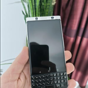 以下が含まれることがあります： 黒い物理キーボードが付いた銀色のスマートフォン。キーボードには「QWERTYUIOPASDFGHJKLZXCVBNM」の文字が刻まれています。スマートフォンは手で持たれています。