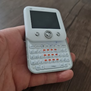 Op de afbeelding: Witte mobiele telefoon met een QWERTY-toetsenbord en een klein scherm. De telefoon heeft een zilveren rand rond het scherm en een zilveren knop aan de voorkant.