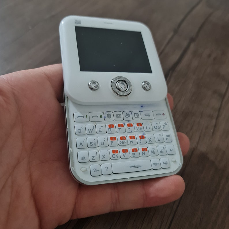 Op de afbeelding: Witte mobiele telefoon met een QWERTY-toetsenbord en een klein scherm. De telefoon heeft een zilveren rand rond het scherm en een zilveren knop aan de voorkant.