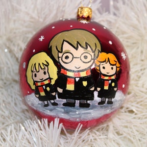 Puede incluir: Una bola de Navidad roja con una ilustración pintada a mano de tres personajes de dibujos animados. Los personajes visten túnicas negras y bufandas con rayas rojas y amarillas. La bola tiene una tapa dorada y está decorada con copos de nieve blancos.