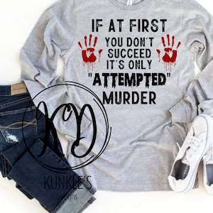 May include: A light gray long-sleeve shirt with the text "IF AT FIRST YOU DON'T SUCCEED IT'S ONLY 'ATTEMPTED' MURDER". The shirt features two red handprints with dripping blood. Dark blue jeans and white sneakers are also visible.