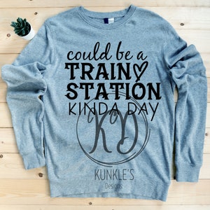 Può includere: Una felpa di colore blu chiaro con il testo "could be a train station kinda day" e un cerchio nero con la lettera "K" al suo interno. Il testo è in nero e il cerchio è grigio. La felpa è su una superficie di legno.