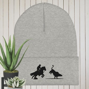 May include: Gray knit beanie with a black embroidered silhouette of a cowboy riding a bucking horse and a bull.
