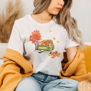 May include: White t-shirt with a green pickup truck filled with pumpkins, gourds, and fall foliage. The truck has the word "MAINE" on the back. A red and orange tree is in the background with falling leaves.