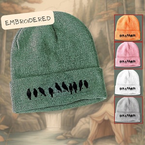 May include: A green knit beanie with a row of black birds embroidered on the brim. The beanie is folded down and has the word "EMBROIDERED" written on a white tag.