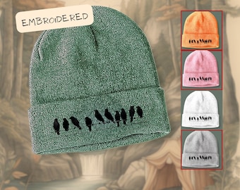 May include: A green knit beanie with a row of black birds embroidered on the brim. The beanie is folded down and has the word "EMBROIDERED" written on a white tag.