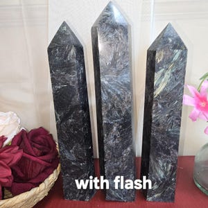 May include: Three dark gray crystal obelisks with pointed tops. The crystals have a textured, reflective surface with streaks of silver and green. The text "with flash" is at the bottom of the image.