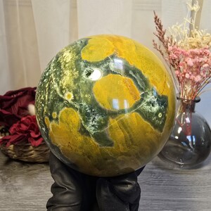 May include: A polished spherical stone featuring a marbled pattern of deep forest green and mustard yellow. The surface displays natural inclusions and crystalline textures, with a smooth, reflective finish that catches light across its rounded form.
