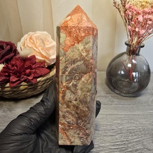 May include: A polished stone tower with a hexagonal shape, featuring a marbled pattern of deep reddish-brown and muted sage green tones. The surface displays natural veining and crystalline textures throughout the vertical, multi-faceted structure.
