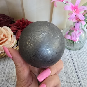 May include: A dark gray sphere with a textured surface is held in a hand with pink nail polish. The sphere is the focal point, with blurred flowers and a vase in the background. The sphere appears to be made of metal or stone.