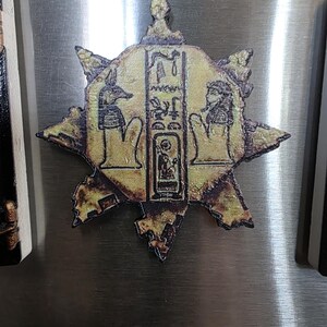 Puede incluir: Un im&aacute;n dorado en forma de estrella con un dise&ntilde;o jerogl&iacute;fico egipcio. El im&aacute;n presenta im&aacute;genes de Anubis y otras figuras, con detalles intrincados y un acabado envejecido. El im&aacute;n probablemente est&aacute; hecho de metal o un material similar.