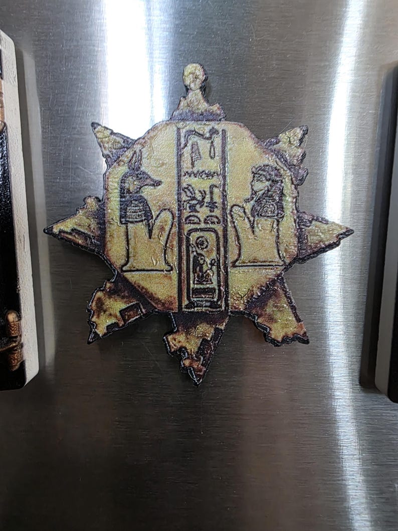 Puede incluir: Un im&aacute;n dorado en forma de estrella con un dise&ntilde;o jerogl&iacute;fico egipcio. El im&aacute;n presenta im&aacute;genes de Anubis y otras figuras, con detalles intrincados y un acabado envejecido. El im&aacute;n probablemente est&aacute; hecho de metal o un material similar.