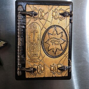 Puede incluir: Un libro rectangular decorativo con borde negro y cubierta dorada. La portada presenta un s&iacute;mbolo Ankh, jerogl&iacute;ficos y un dise&ntilde;o de estrella con un ojo. El libro tiene cierres met&aacute;licos y un lomo con l&iacute;neas verticales.