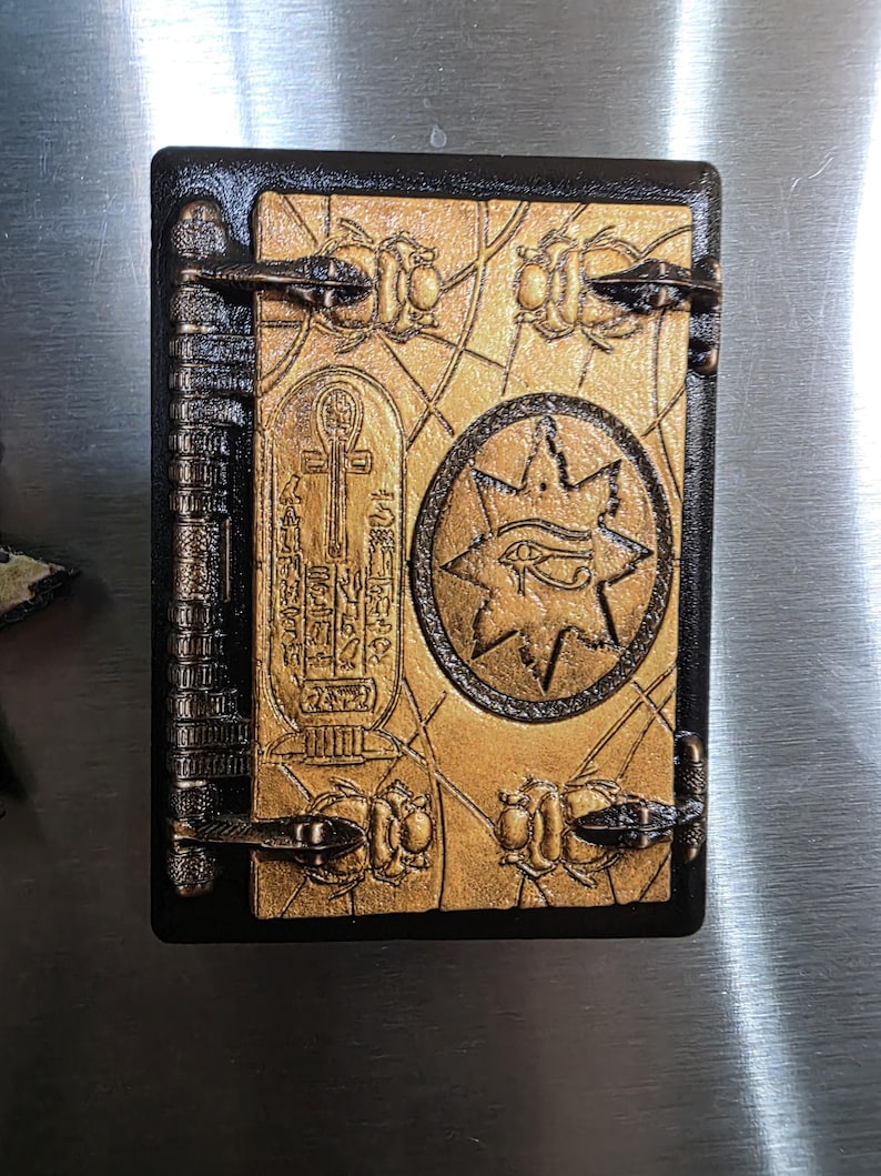 Puede incluir: Un libro rectangular decorativo con borde negro y cubierta dorada. La portada presenta un s&iacute;mbolo Ankh, jerogl&iacute;ficos y un dise&ntilde;o de estrella con un ojo. El libro tiene cierres met&aacute;licos y un lomo con l&iacute;neas verticales.