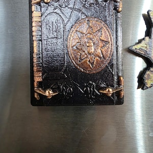 Puede incluir: Un im&aacute;n negro con forma de libro de cuero sint&eacute;tico con detalles en color bronce. El im&aacute;n presenta un emblema circular con un escarabajo y un dise&ntilde;o de estrella. El texto "NO HARM EVER CAME FROM READING A BOOK" est&aacute; en la parte superior.