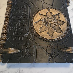 May include: A large, dark gray book with gold accents, featuring Egyptian hieroglyphs and a central emblem of a scarab beetle within a starburst design. The book's cover is intricately detailed with embossed patterns and insect motifs, suggesting an ancient or fantasy theme.