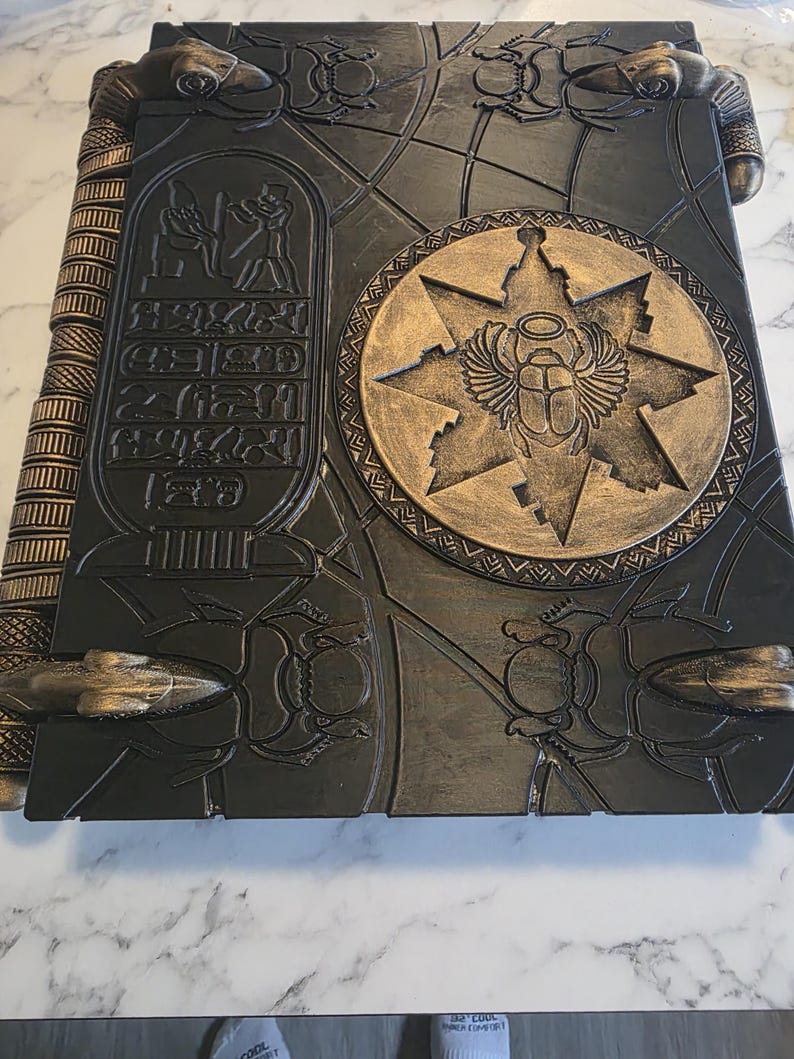 May include: A large, dark gray book with gold accents, featuring Egyptian hieroglyphs and a central emblem of a scarab beetle within a starburst design. The book's cover is intricately detailed with embossed patterns and insect motifs, suggesting an ancient or fantasy theme.