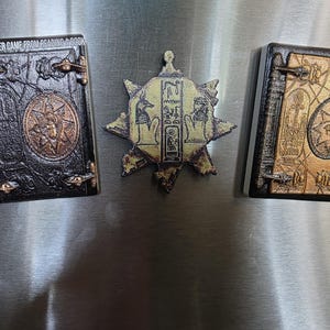 Puede incluir: Tres imanes decorativos con tem&aacute;tica del antiguo Egipto. Dos tienen forma de libro con dise&ntilde;os intrincados y cierres met&aacute;licos. El tercero tiene forma de estrella con jerogl&iacute;ficos. Un im&aacute;n con forma de libro tiene el texto "NO HARM EVER CAME FROM READING A BOOK".