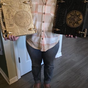May include: Two decorative books with intricate designs. One is gold-colored, the other black. Both have detailed covers with Egyptian-style hieroglyphs and symbols. The books are held by a person, showcasing their size and craftsmanship. The covers have a raised, textured appearance.