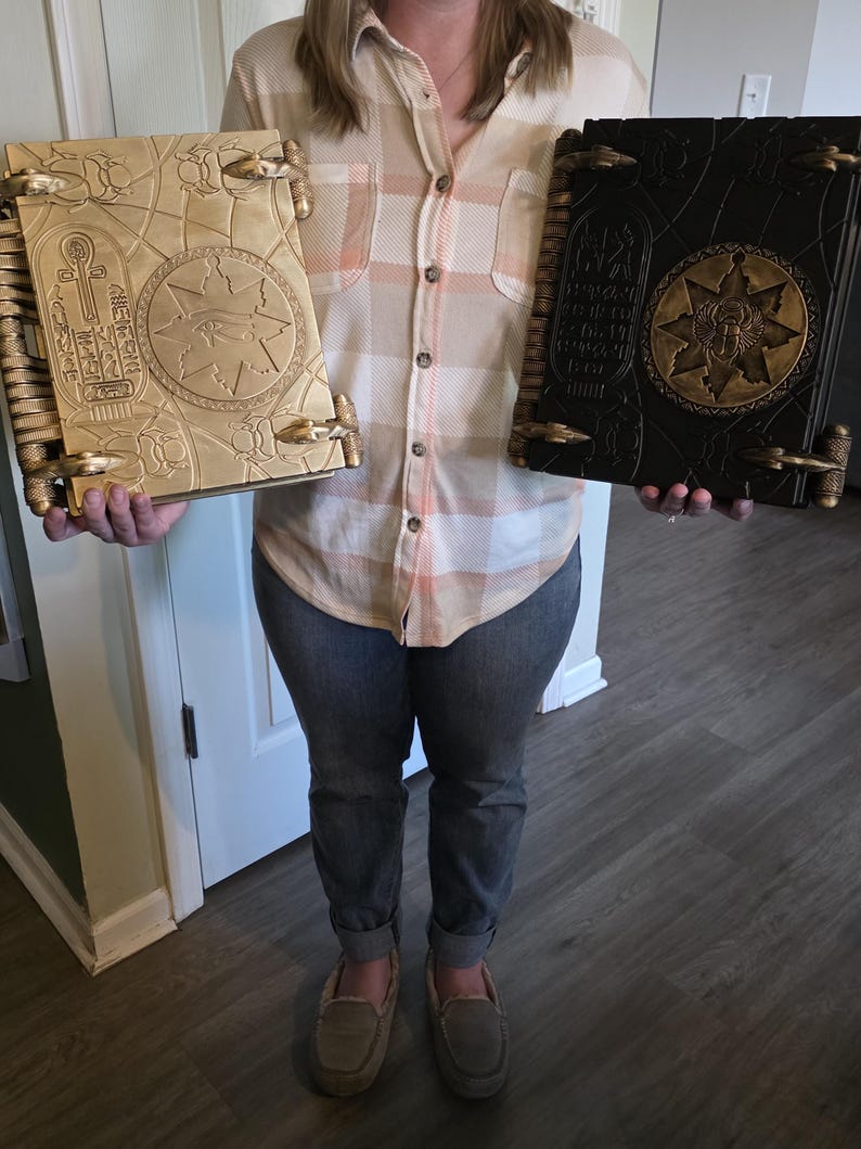 May include: Two decorative books with intricate designs. One is gold-colored, the other black. Both have detailed covers with Egyptian-style hieroglyphs and symbols. The books are held by a person, showcasing their size and craftsmanship. The covers have a raised, textured appearance.